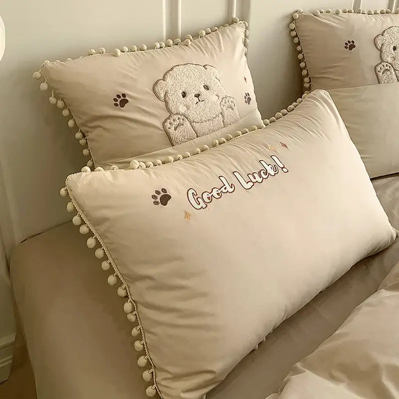 Cream-colored cotton bedding set pillow with embroidered cartoon bear applique and good luck text trimmed in pom-poms