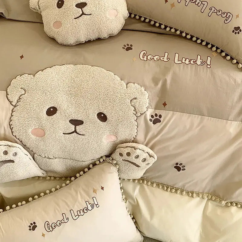 Cream-colored plush teddy bear pillow with embroidered facial features and pink cheeks in cotton bedding set with cartoon bear applique