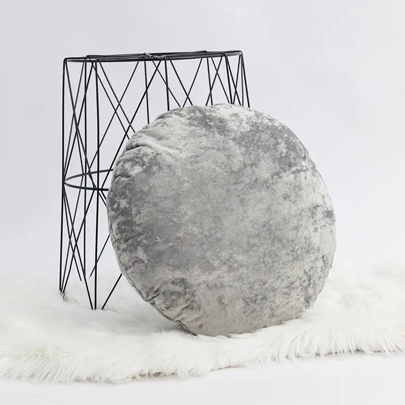 A plush round gray velvet pillow cover with a dark grain texture on a white faux fur rug for living room decor