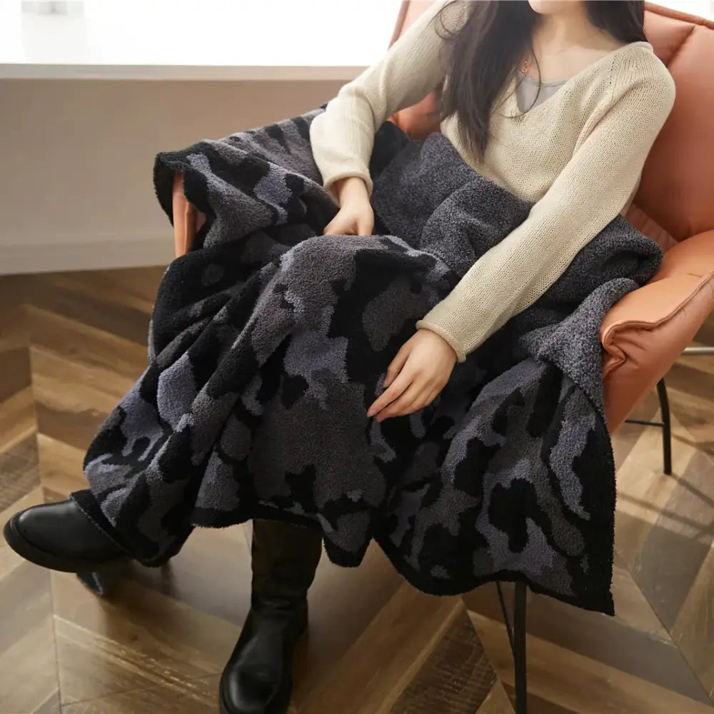 A plush dark gray and black camouflage knitted throw blanket draped over a person’s lap for cozy use