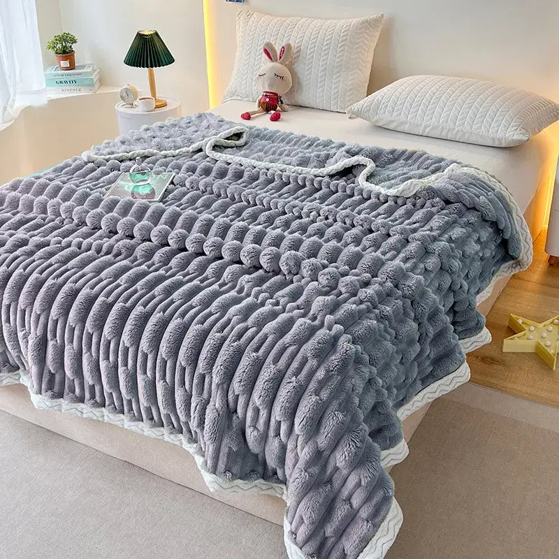 A plush gray knitted throw blanket with pom-poms and white trim styled alongside a pink bamboo velvet blanket with braided trim