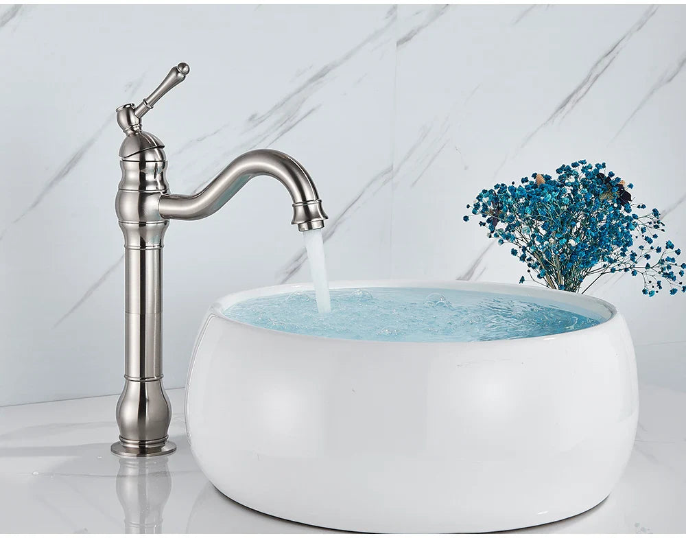 A deck mounted single hole brushed nickel faucet with curved spout and ornate detailing beside a white vessel sink