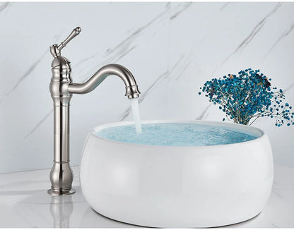A deck mounted single hole brushed nickel faucet with curved spout and ornate detailing beside a white vessel sink