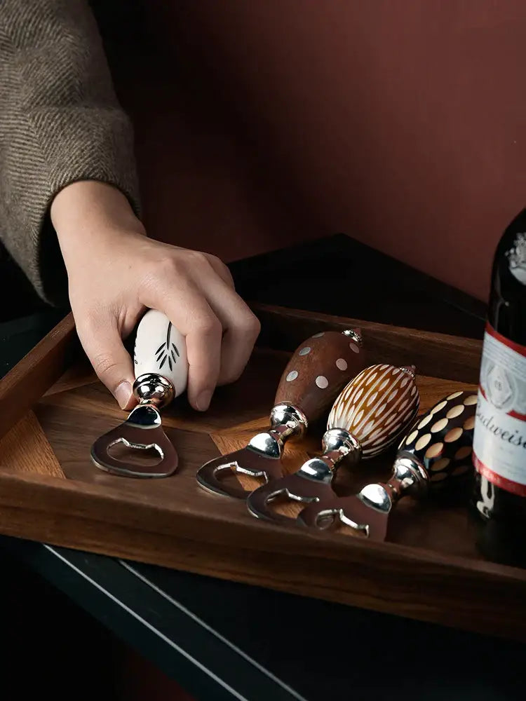 A hand holds a white ceramic beer bottle opener with black leaf etching and a polished silver metal cap