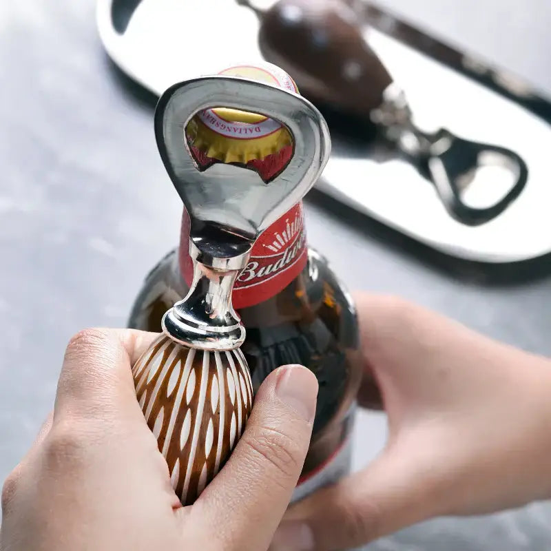 A sleek silver beer bottle opener with brown and white striped wooden handle opening a Budweiser beer bottle for enhanced drinking experience