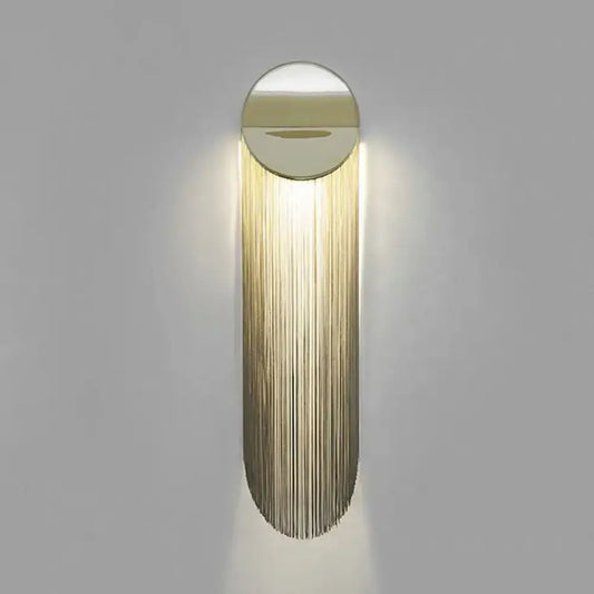 Postmodern gold fringed wall light fixture with sleek oval design and cascading metal strands emits warm light for bedroom