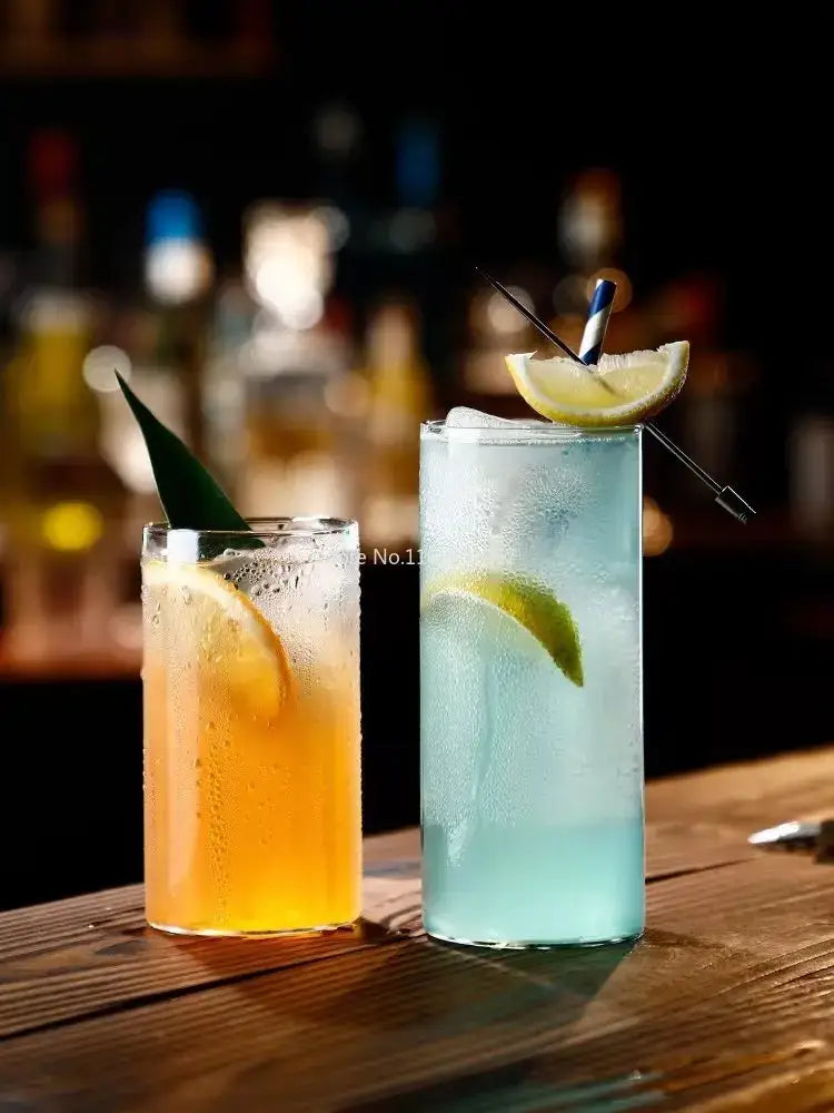 Two tall clear glass highball cups with amber and light blue cocktails, ice cubes, citrus garnish, and straws in a premium set