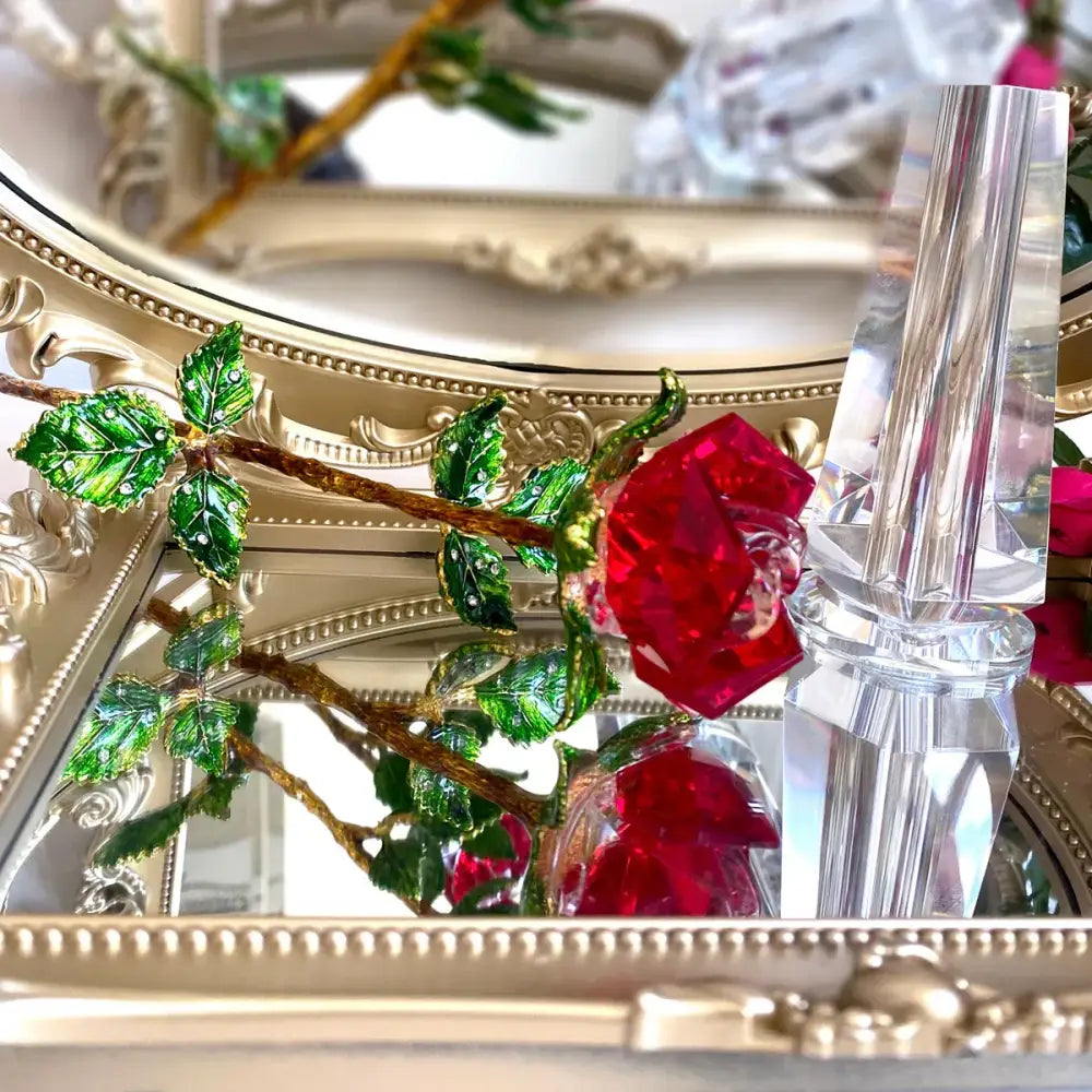 A sparkling red crystal rose figurine with green enamel leaves and gold-toned stem displayed on an ornate mirror surface