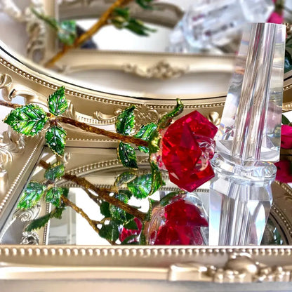A sparkling red crystal rose figurine with green enamel leaves and gold-toned stem displayed on an ornate mirror surface