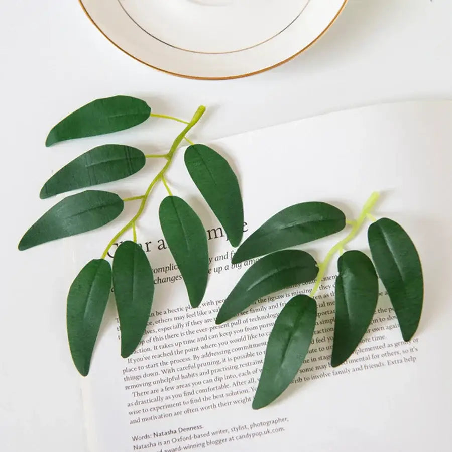 A branch of dark green silk willow leaves with glossy finish and light green stems arranged on printed paper