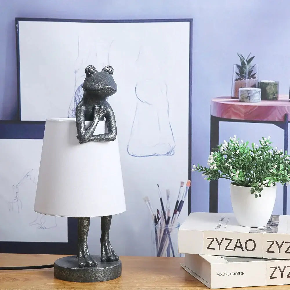 A dark metallic decorbites frog resin desk lamp with crossed arms base and white lampshade for bedroom lighting