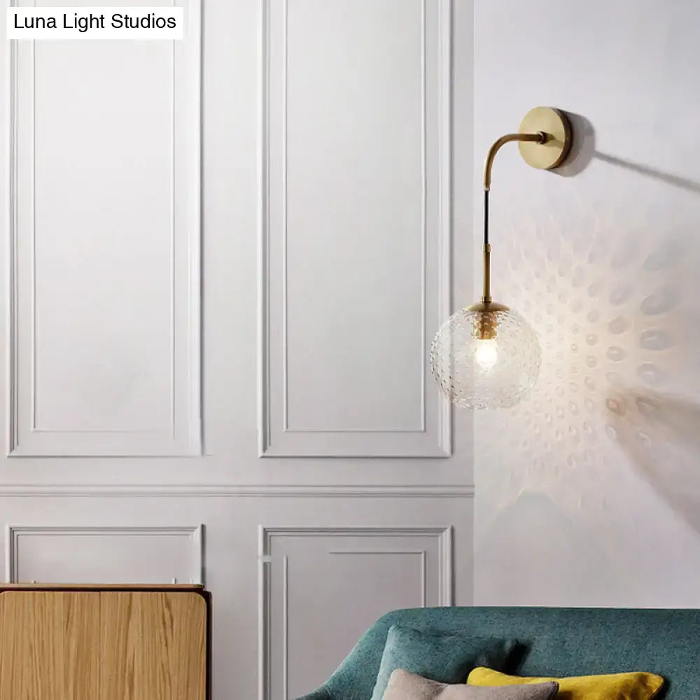 A brass-finished wall sconce with a curved arm and clear globe bulb for retro pink yellow blue bedroom lighting