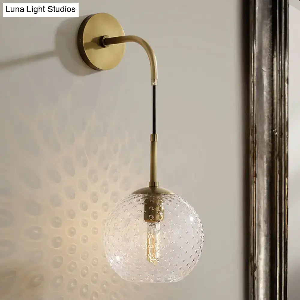 A brass wall sconce with curved arm and clear textured glass globe for pink yellow blue retro bedroom lighting