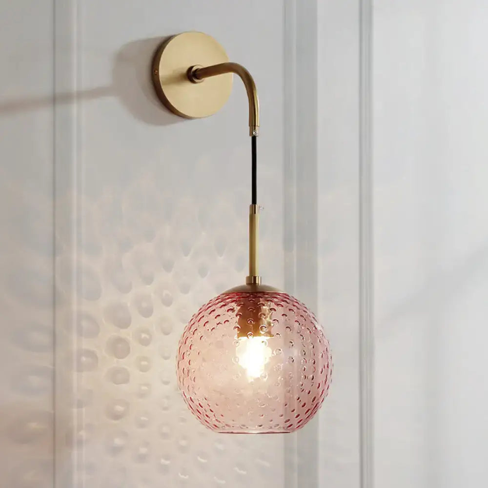 A pink glass globe wall lamp with hammered texture suspended by brass arm and black cord in pink yellow blue style