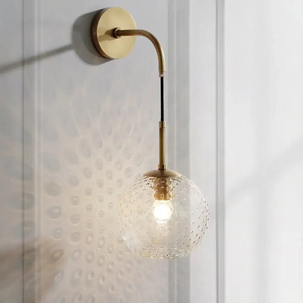 Gold-toned wall sconce with curved arm and textured clear glass globe for pink yellow blue retro bedroom lighting