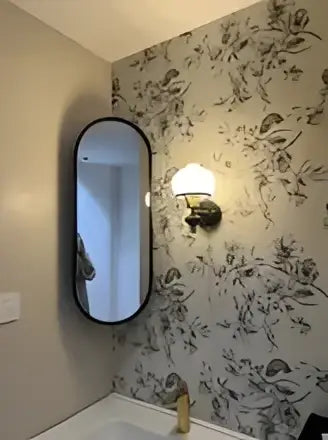 Oval-shaped mirror with sleek black frame reflecting floral wallpaper paired with retro brass and black french wall lamp decorbites french wall