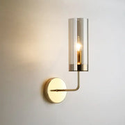 A sleek brass wall mount lamp with a cylindrical smoked glass shade and exposed filament bulb for living room use