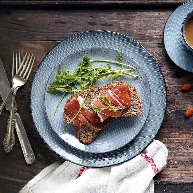 Blue ceramic dinner plate with vertical ridges and speckled glaze holding dark bread with prosciutto and arugula decorbites ceramic dinner