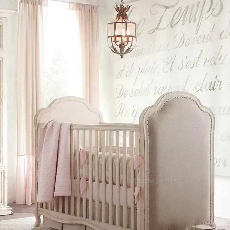 A soft beige upholstered crib with curved headboard and pink accents paired with decorbites grey gold metal cage chandelier