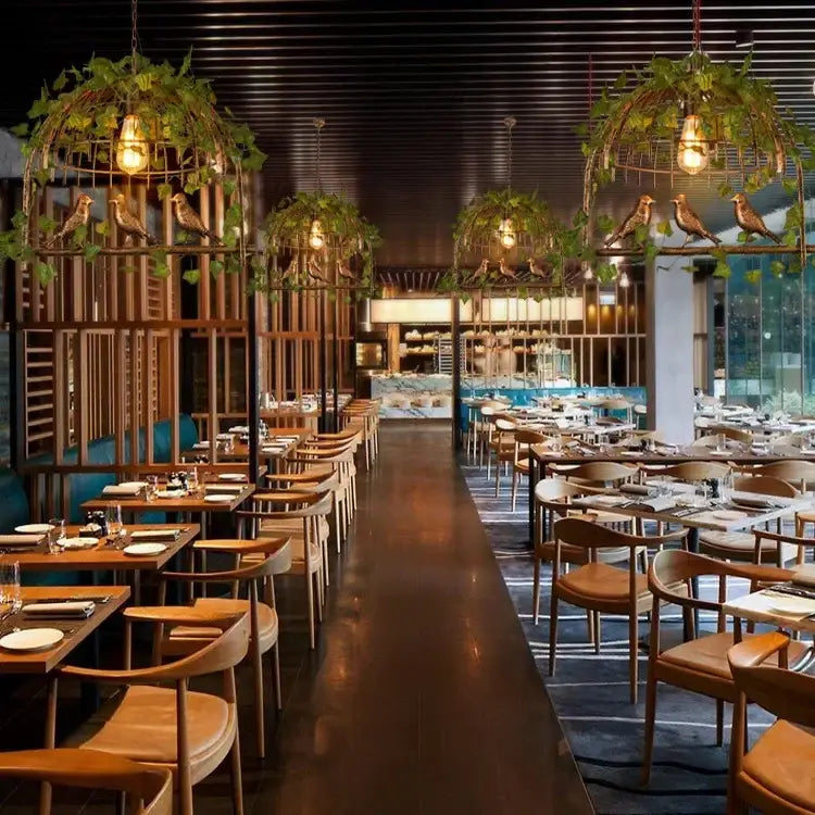 Rustic bronze iron bird cage chandelier with greenery above wooden tables and chairs with brown leather seats in restaurant