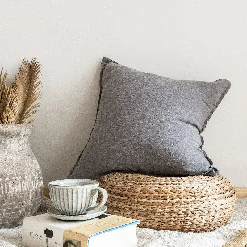 Gray textured linen fringed pillow cover resting on woven wicker mat for rustic earthy tones living room decorbites decor