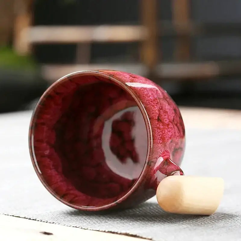 A glossy crimson ceramic latte mug with textured swirling glaze and a smooth wooden handle for rustic drinkware