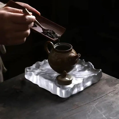 Earthenware teapot with rounded body and short spout on ornate silver tray beside scalloped crystal glass coaster and jewelry display panel