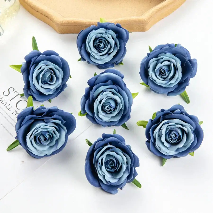 Six silk blue artificial flowers with layered petals and green stems arranged on white surface for wedding decor