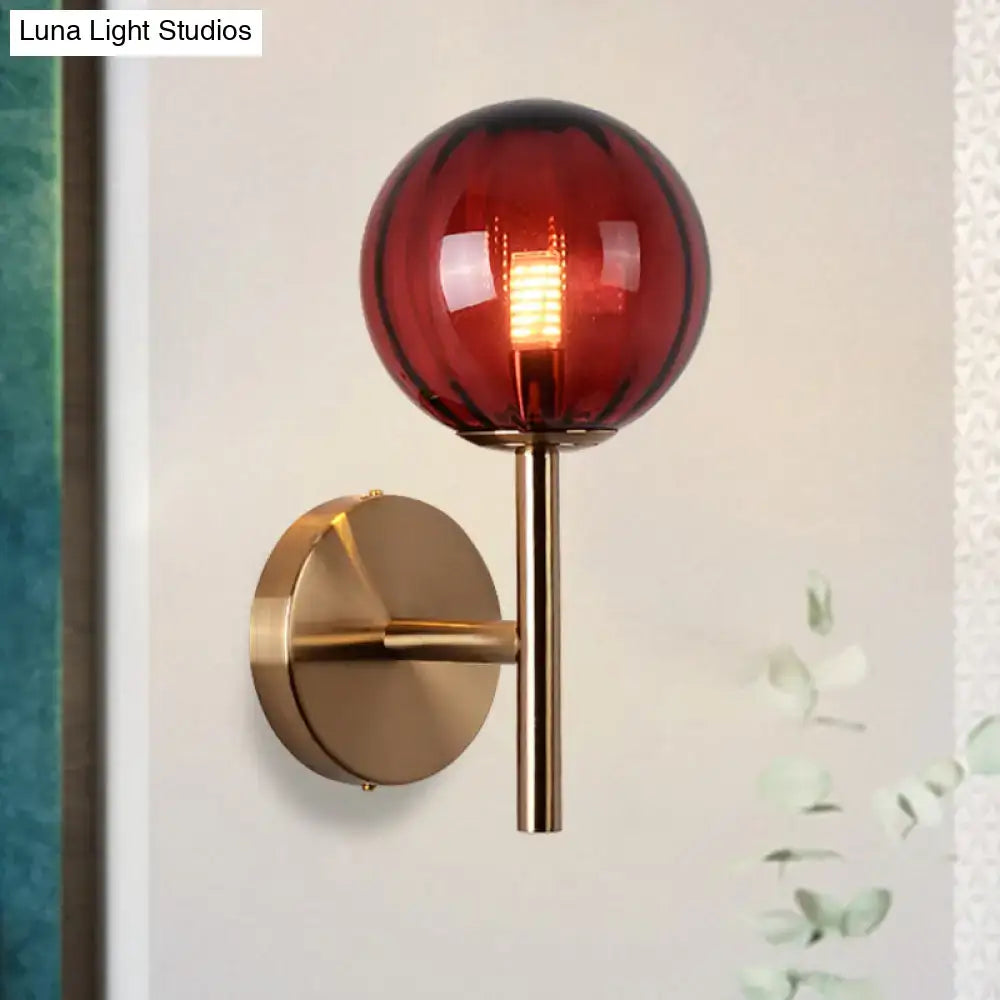 A sleek brass bedside sconce light with a deep red spherical glass globe featuring subtle vertical ridges for round wall lighting in red green amber