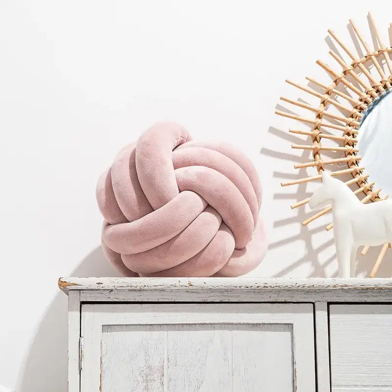 A soft pink velvet knotted ball round cushion with plush texture resting on a white wooden cabinet for bedroom decor
