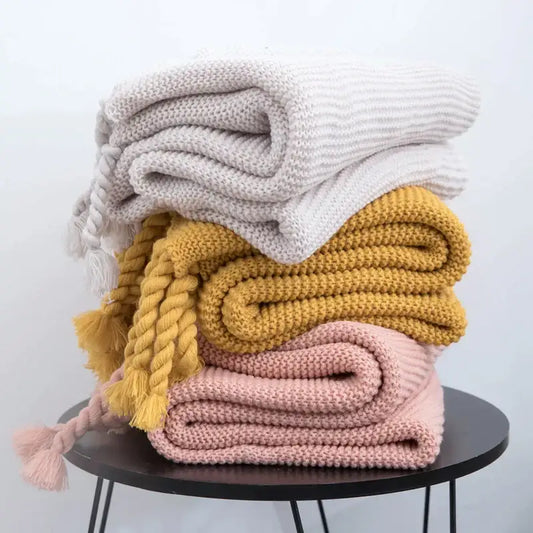 Stack of three decorbites knit throw blankets in ivory mustard yellow and blush pink with ribbed texture and braided tassels