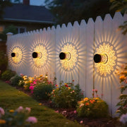 Four solar-powered datura lantern wall lamps with golden filigree cast warm sunburst patterns on white picket fence