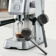 Silver stainless steel solis espresso machine brewing dark coffee with black handle alongside stainless steel coffee maker brush