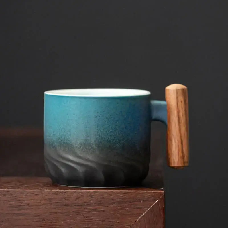 A ceramic coffee cup with teal gradient glaze and wood handle resting on a dark wood surface for kitchen use