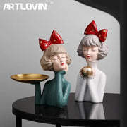 Two stylized bowknot girl resin figurines in teal and white with red bows holding a gold tray and golden apple decorbites bowknot girl