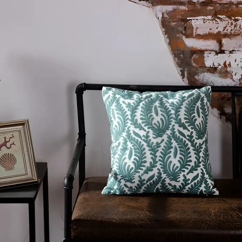Teal and white blue geometric embroidered cushion cover with leafy damask design resting on dark brown leather bench decorbites
