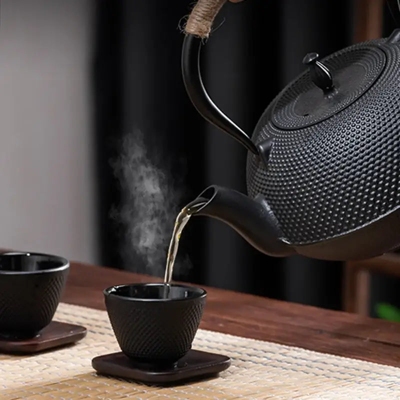 Textured black cast iron tea pot with woven handle pouring steaming tea into matching black cast iron tea cups
