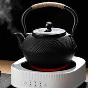 Black cast iron tea pot with textured surface and woven handle on white electric kettle base for kitchen steeping