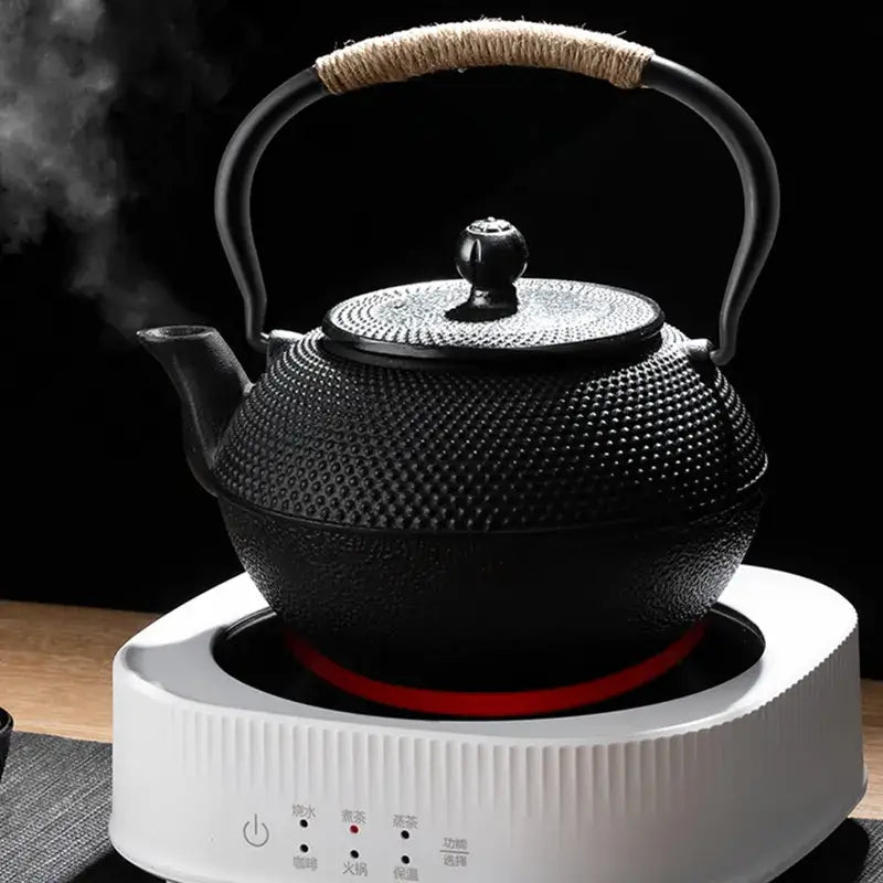 Black cast iron tea pot with textured surface and woven handle on white electric kettle base for kitchen steeping