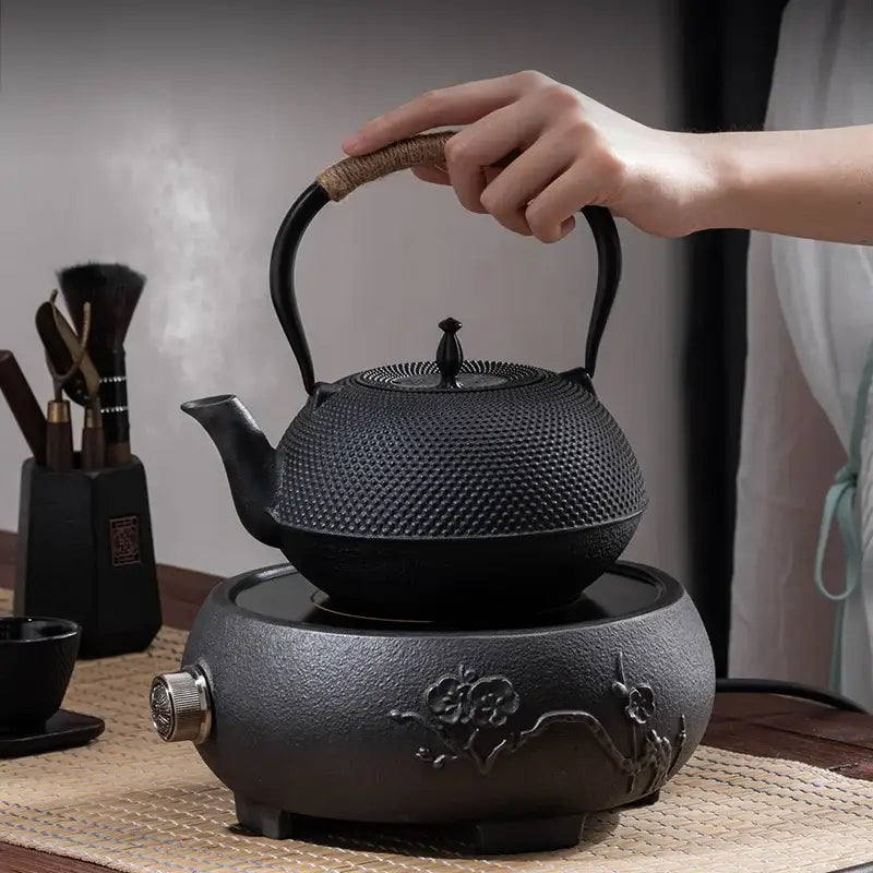 Textured black cast iron tea pot with woven handle resting on matching electric heating base for kitchen steeping