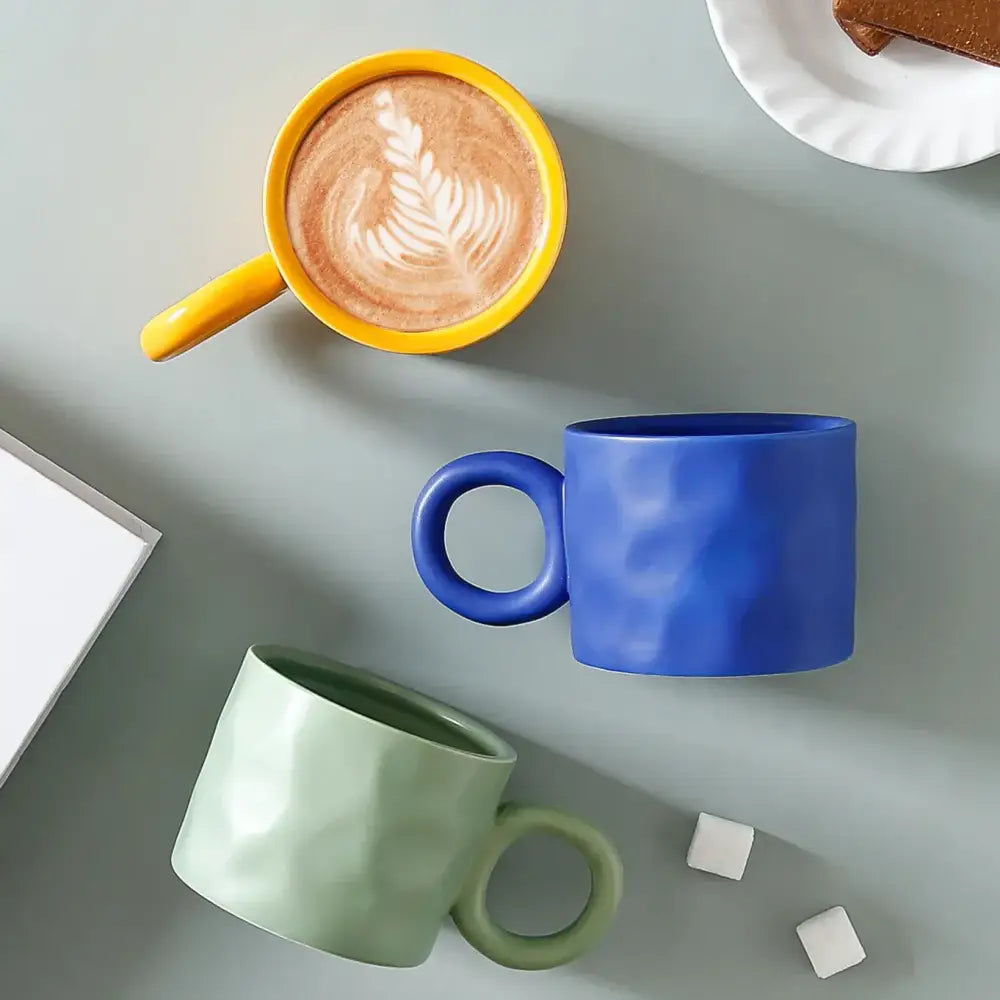Three ceramic coffee mugs in vibrant yellow, deep blue, and soft green with textured surfaces and round handles