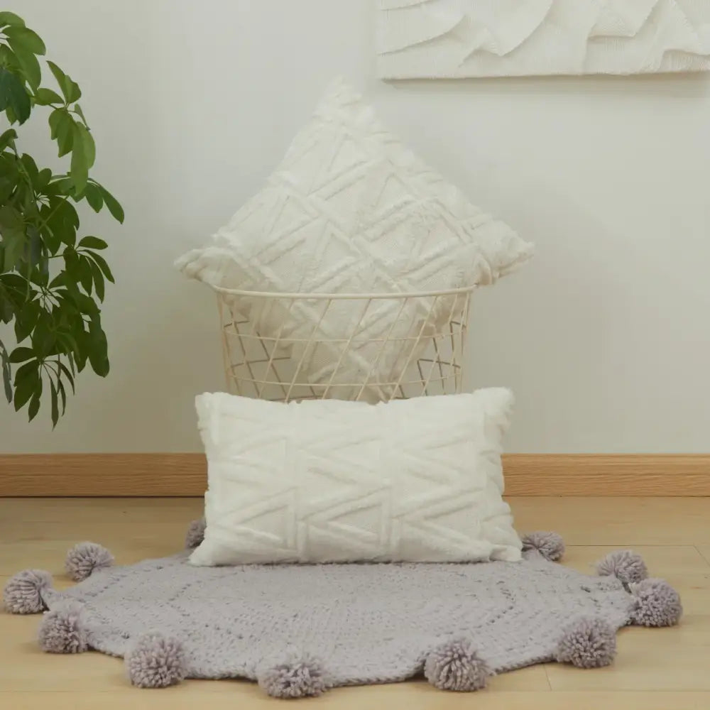 A soft light gray knitted rug with fluffy pom-poms complements a textured earthy geometric diamond embroidery living room cushion cover