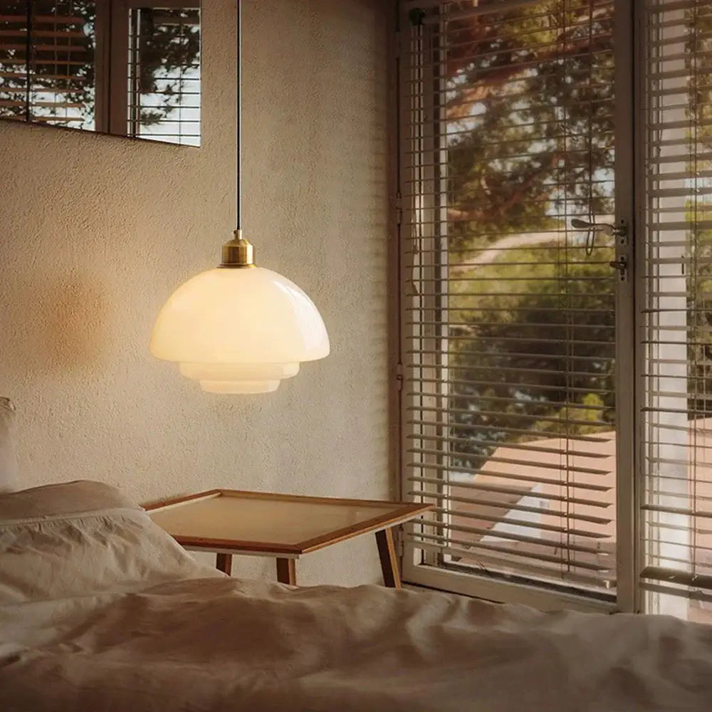 A tiered cream glass pendant light with layered opal shade and brass hardware hangs above a bedside table