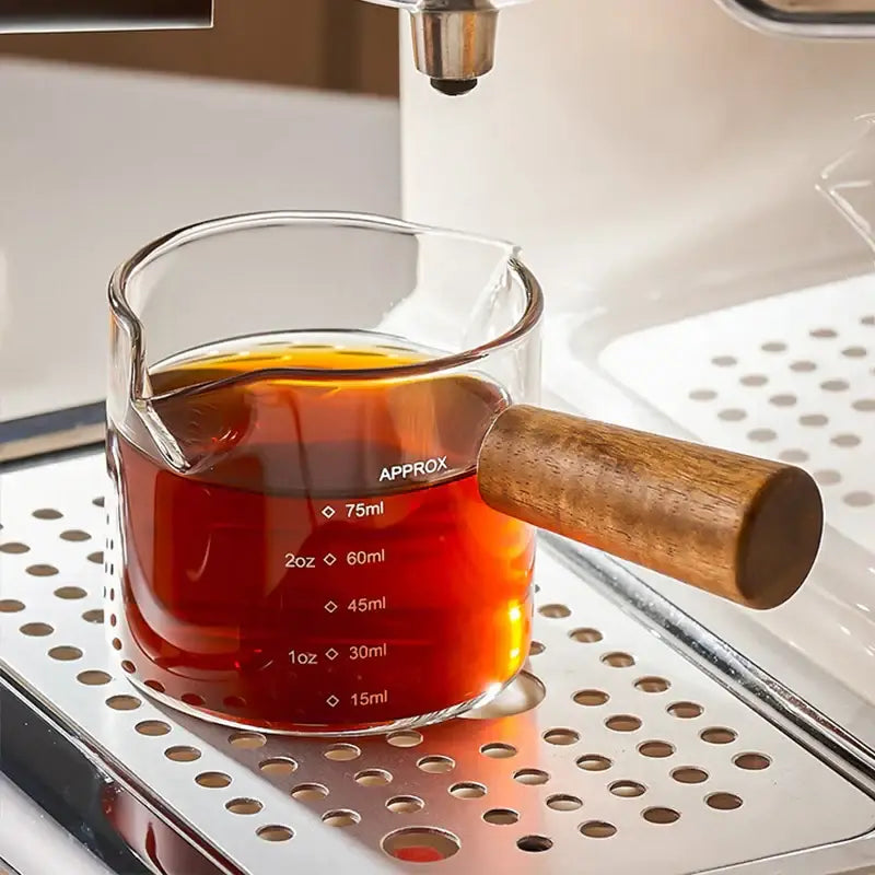 Transparent glass espresso measuring cup with wood handle filled with amber liquid and marked with ounces and milliliters