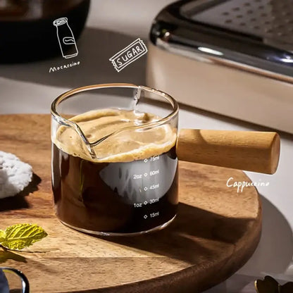 A transparent glass espresso measuring cup with wood handle filled with dark coffee topped with creamy foam