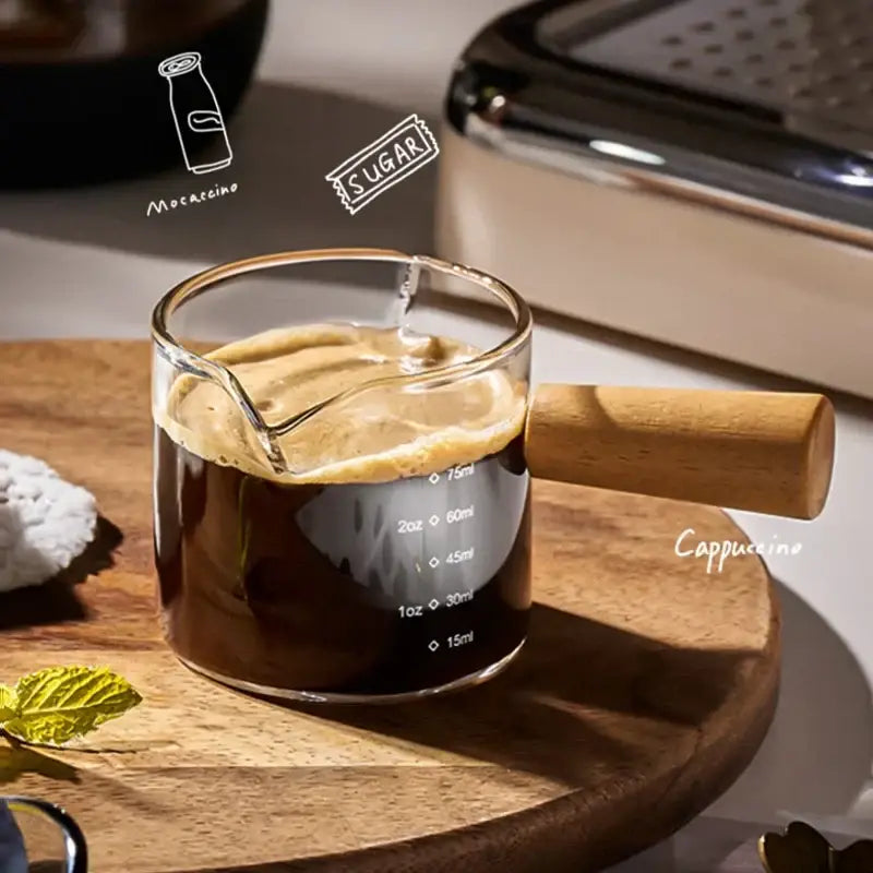 A transparent glass espresso measuring cup with wood handle filled with dark coffee topped with creamy foam