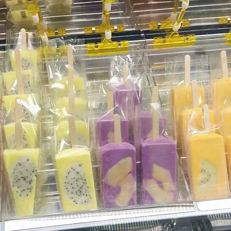 Transparent pink ice cream bags with gold ribbon containing yellow and purple fruit-flavored popsicles with dragonfruit seeds and mango slices