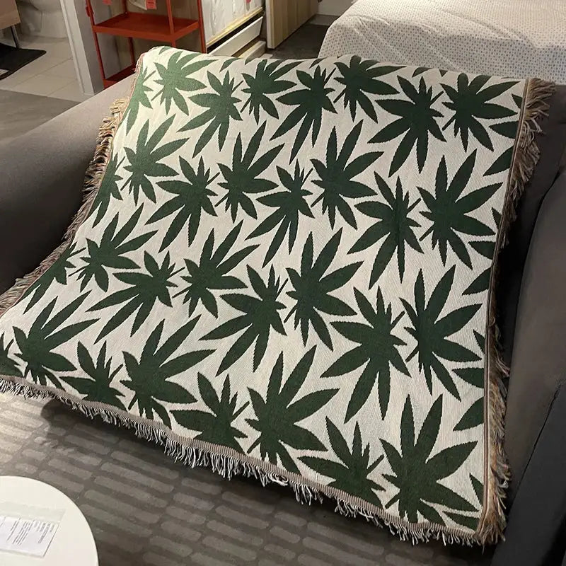 A cozy knit throw blanket in cream with dark green cannabis leaf pattern and soft frayed edges for decorbites cozy knit decor
