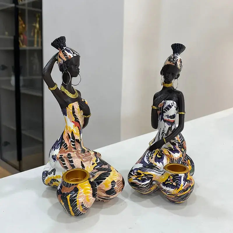 Two vibrant resin decorbites black woman figurines in patterned dresses with gold jewelry beside a black woman candlestick