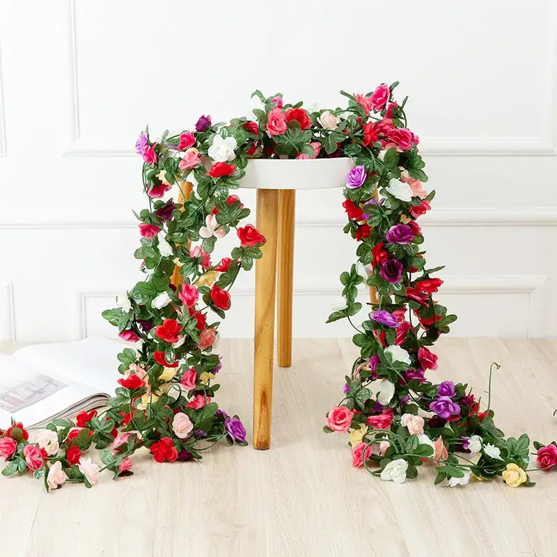 A vibrant multi colored rose flowers vine garland with lush green leaves draped over a wooden stool for living room decor